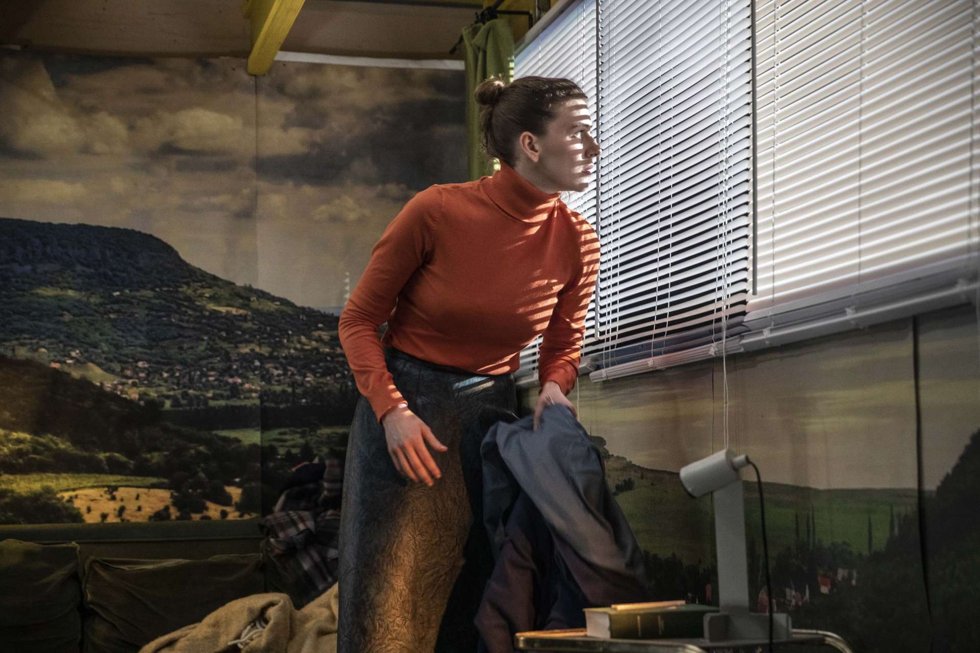Une femme habillée d'un col roulé orange regardent avec inquiétude entre les lattes de store vénitien. L'intérieur semble être celui d'une chambre, mais le papier peint est une photographie d'un paysage rural.
