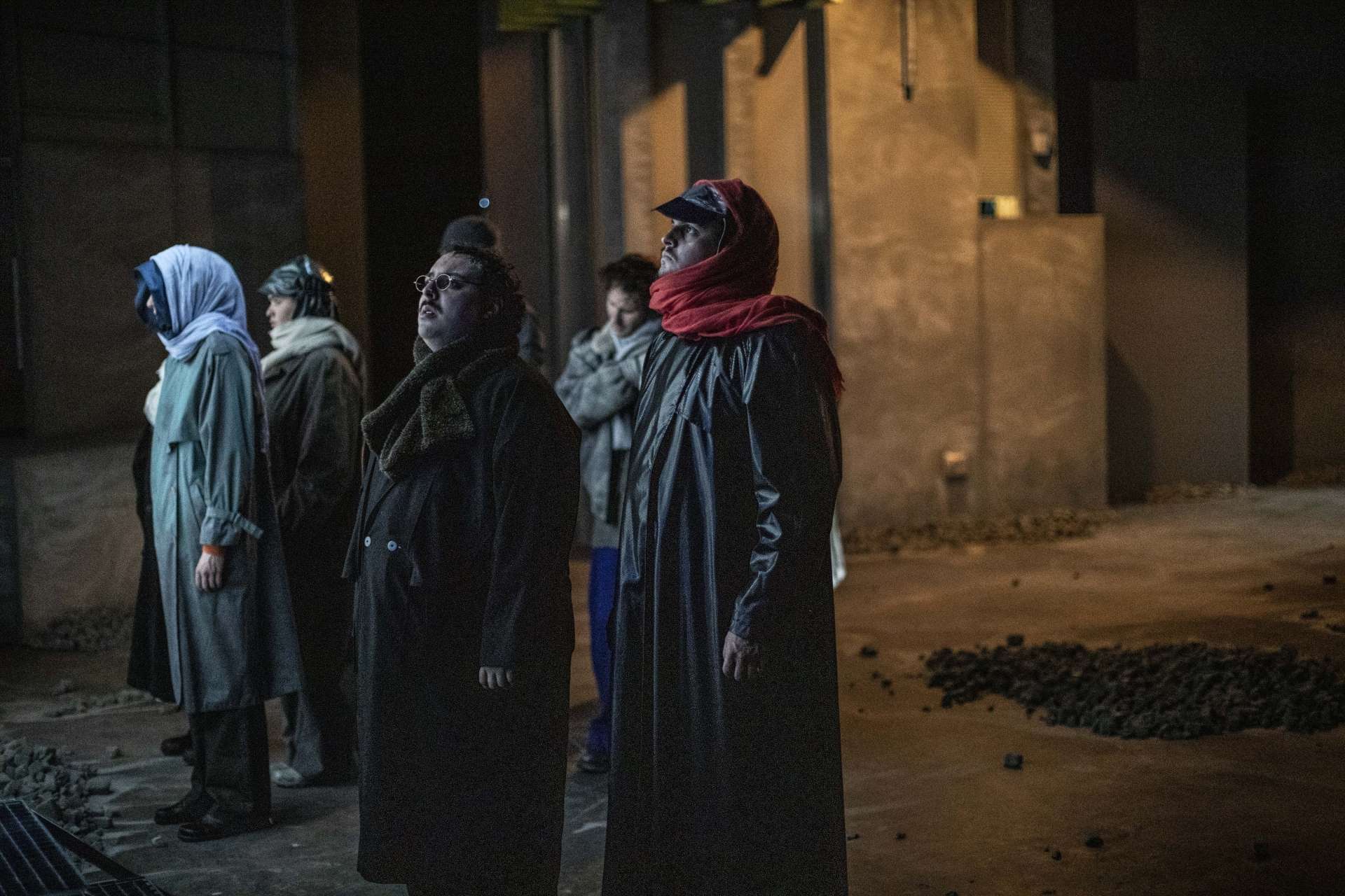 Un groupe de personnes habillées d'imperméables qui couvrent leurs visages, regardent hors champ. Seule une femme regarde et se dirige dans une autre direction.