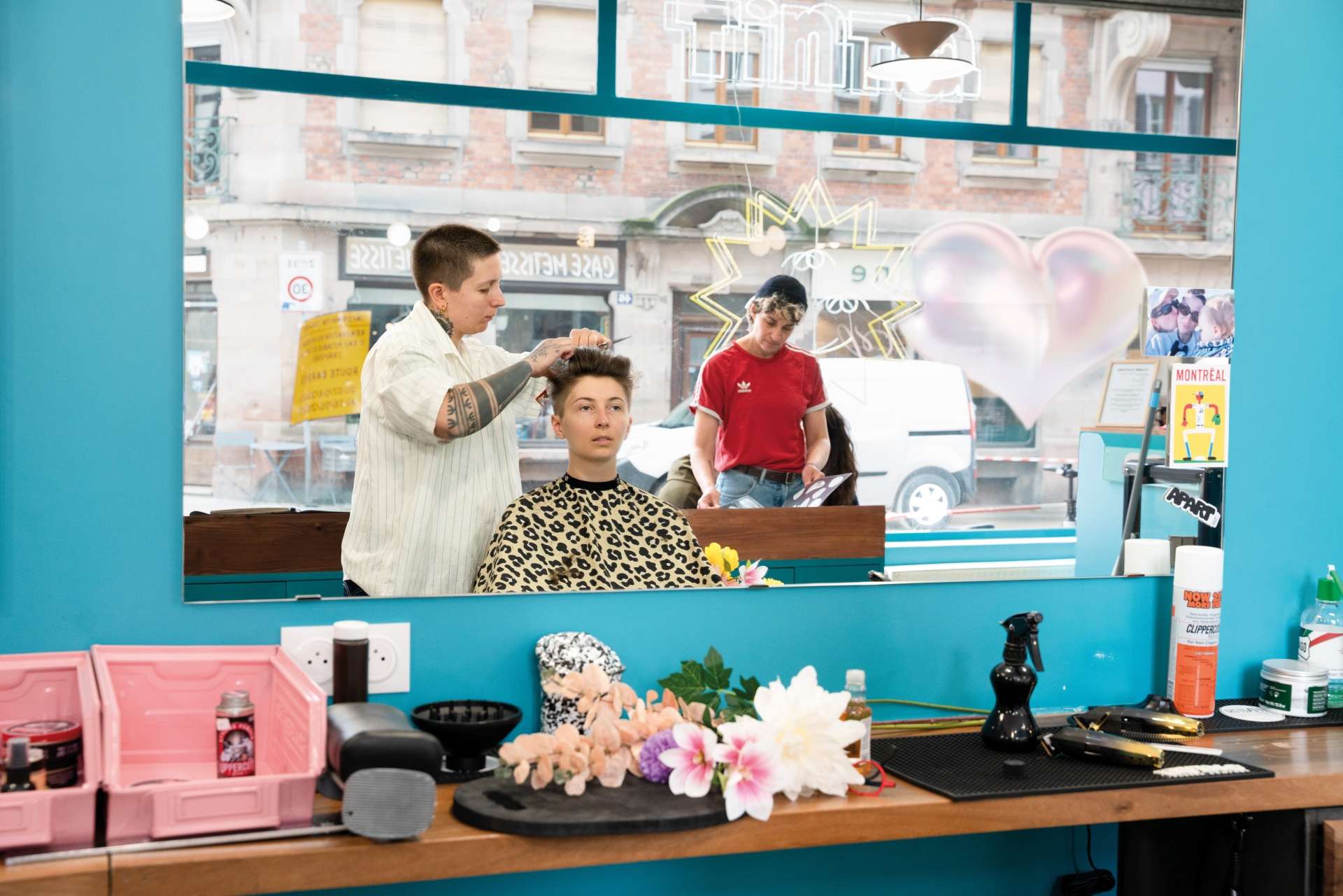 salon de coiffure aux murs bleus 1 femme se fait coiffer par une coiffeuse au premier plan l'aurte au second plan au loin feuillette un magazine 