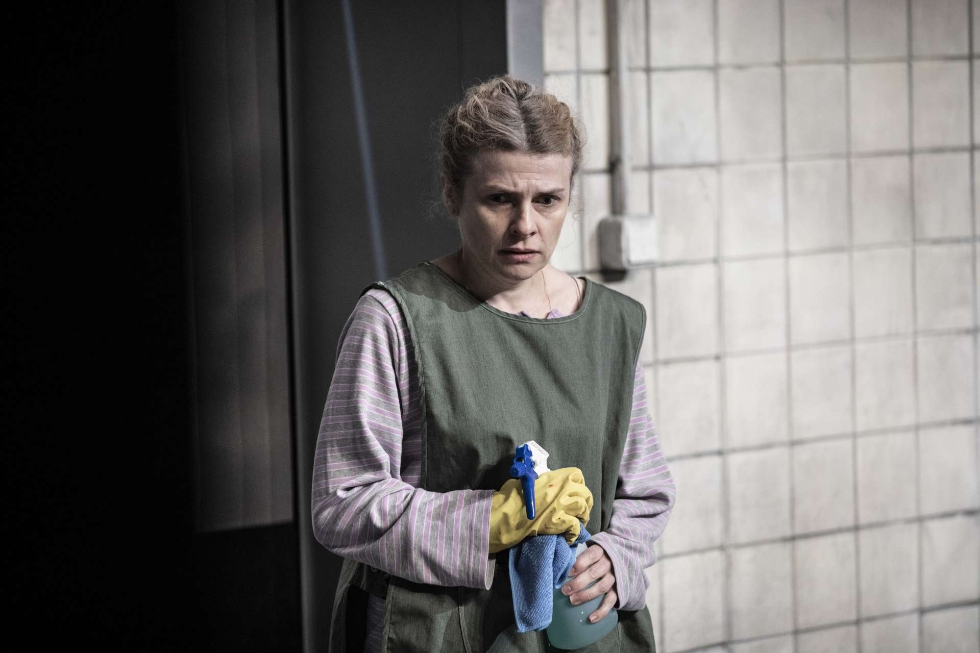 Prendre soin Alexander Zeldin © Jean-Louis Fernandez une actrice seule serre son sac contre elle habillée d'un tablier dans un decor carrelé blanc l'air inquiet