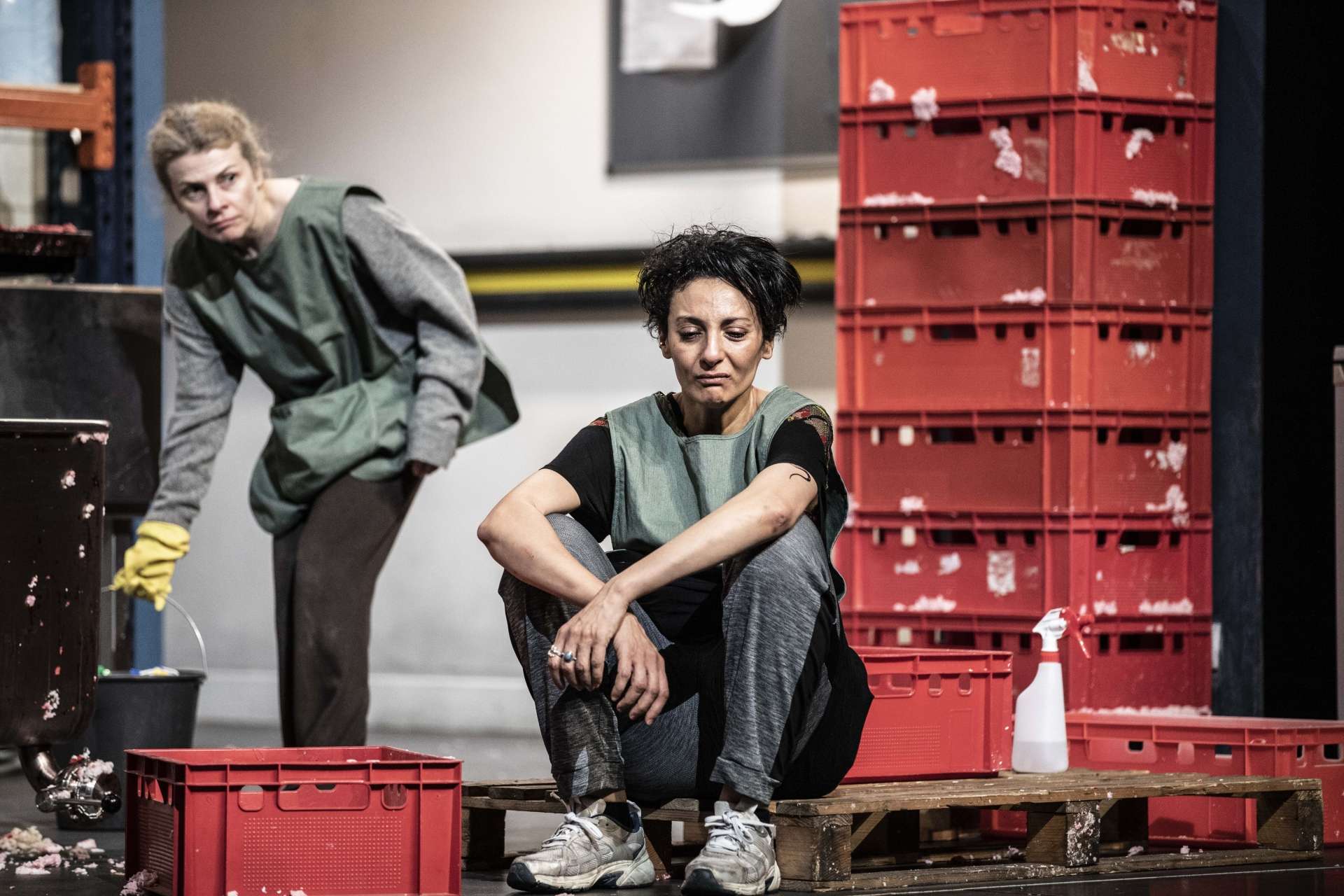 dans un décor interieur industriel deux femmes l'une assise au premier plan silencieuse l'autre debout au second plan la regarde des cagettes rouges les entourent l"une porte des gants jaunes et un seau elles semblent nettoyer le lieu