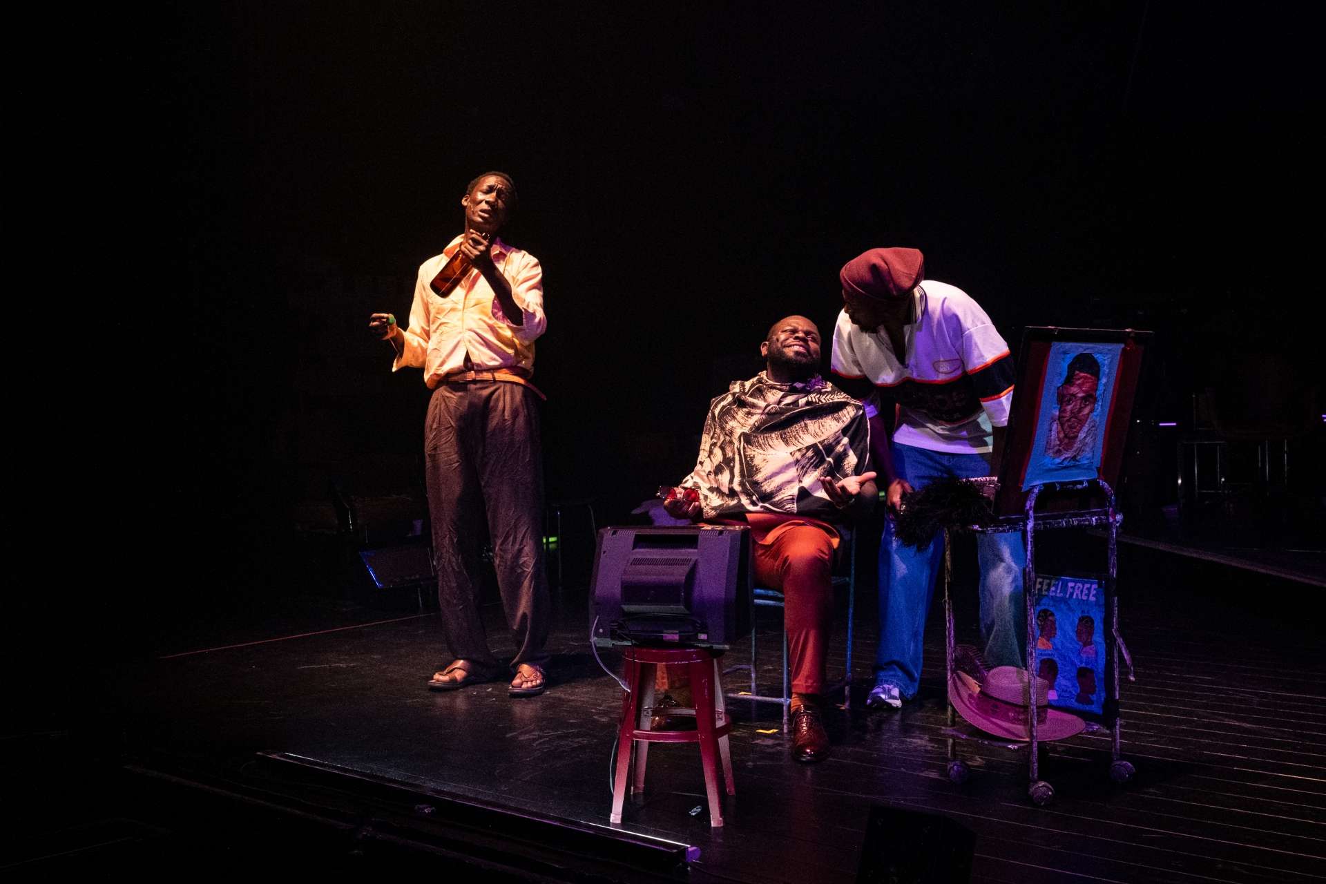 Barber Shop Chronicles © Stef Stessel 3 hommes noirs chez le barbier dont l'un assis, et l'autre portant une bouteille brune à la main